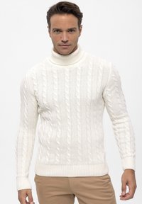 Weißer, kabelgestrickter Pullover mit hohem Stehkragen, gerippten Bündchen und Saum, gestaltet mit strukturierten Mustern. Getragen mit hellbeigen Hosen.