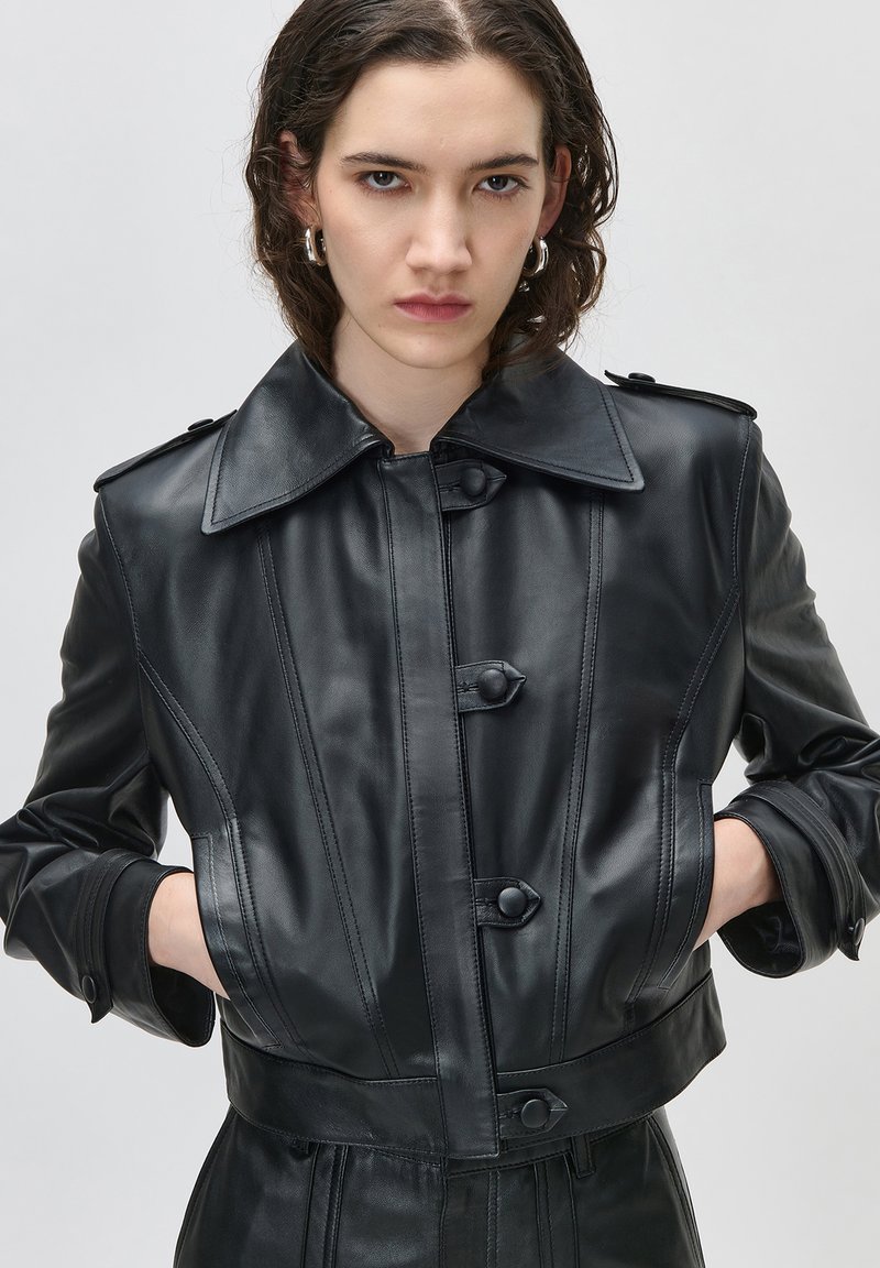 Νεαρός με κοντά σκούρα μαλλιά φορά μαύρο δερμάτινο μπουφάν με μεγάλο γιακά, λεπτομέρειες με κουμπιά και τα χέρια στις τσέπες, πάνω σε απλό φόντο.