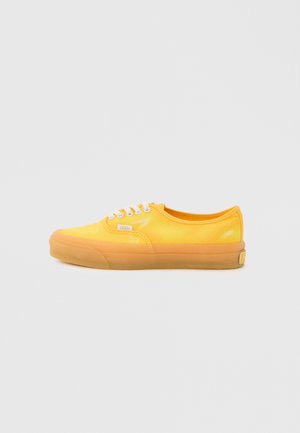 Gele lage Vans-sneaker met witte veters en een rubberen zool, vanaf de zijkant getoond op een lichtgrijze achtergrond.