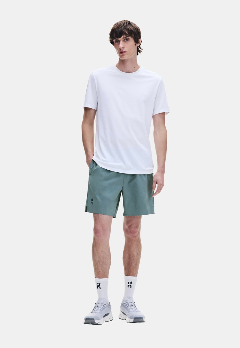 Jeune homme debout portant un t-shirt blanc à manches courtes, un short vert, des chaussettes blanches montantes et des chaussures de sport grises sur un fond uni.