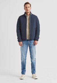 Marineblaue Bomberjacke mit Stehkragen, Reißverschluss und zwei Seitentaschen, getragen über einem hellbeigen Hemd und blauer Jeans, kombiniert mit weißen Turnschuhen.