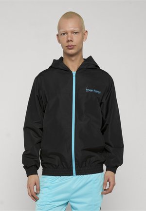 Persona con capelli biondi corti indossa una giacca nera con cappuccio, cerniera e logo blu, abbinata a pantaloni azzurri, in piedi davanti a uno sfondo semplice.