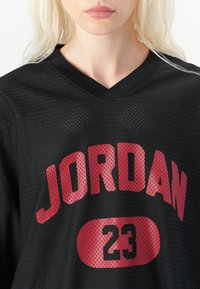 Le maillot en mesh noir arbore l'inscription rouge "JORDAN" et le numéro "23". Il présente un col en V avec un tissu texturé et des perforations respirantes.