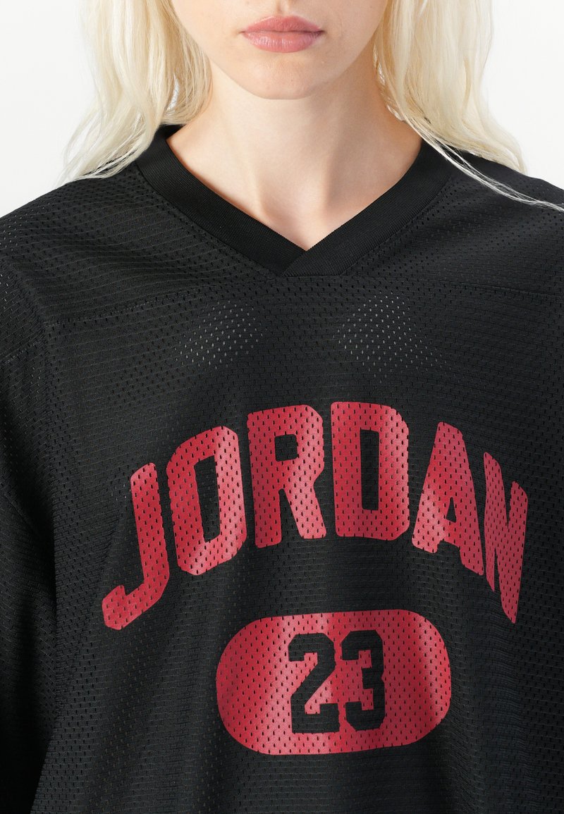 Le maillot en mesh noir arbore l'inscription rouge "JORDAN" et le numéro "23". Il présente un col en V avec un tissu texturé et des perforations respirantes.