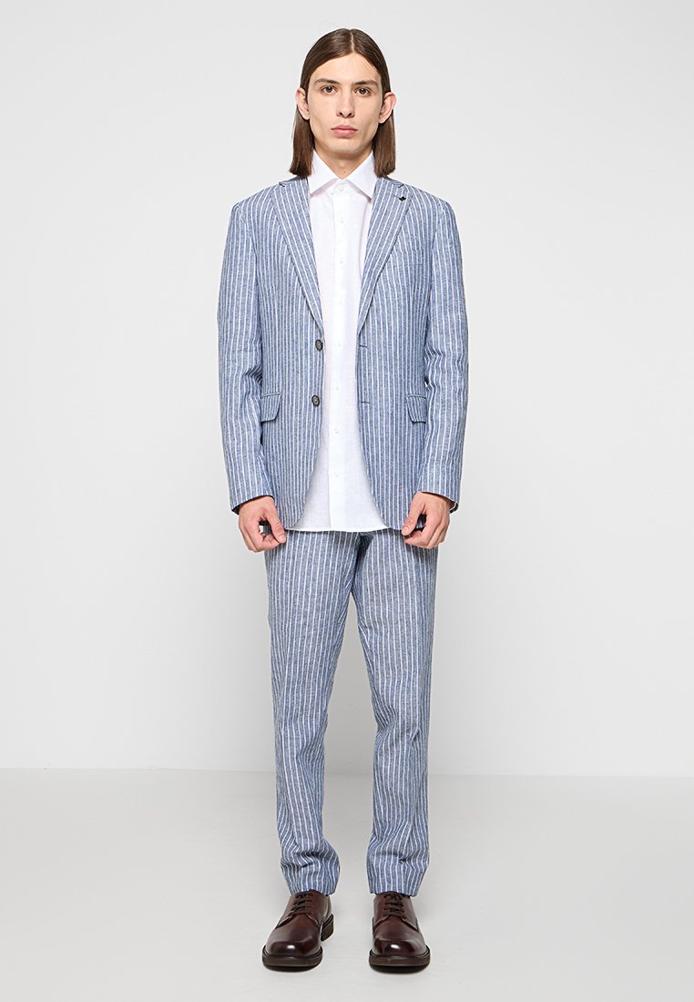 Wielobarwna garsonka w odcieniach niebieskiego i białego, składająca się z dopasowanej marynarki z klapami typu notch, dopasowanych spodni oraz białej koszuli z guzikami. Całość dopełniają brązowe buty.