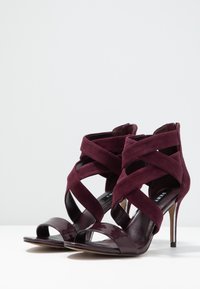 Burgundské lodičky na vysokém podpatku kombinující semiš a lakovanou kůži, s křížovými popruhy a otevřenou špičkou.