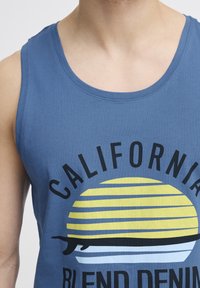 Top de bumbac albastru cu un design grafic reprezentând un soare cu dungi galbene și o siluetă neagră, având text îndrăzneț în negru care spune "CALIFORNIA."