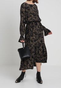 Långärmad klänning med kamouflagemönster i gröna och svarta nyanser, med en avslappnad passform och volangkjol. Kombinerad med svarta ankelstövlar och handväska.