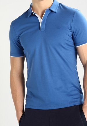 Polo à manches courtes bleu avec un col blanc, une coupe ajustée, et un logo en relief sur la poitrine, fabriqué à partir d'un tissu lisse et léger.