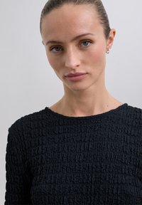 Zwarte top met een gestructureerd oppervlak en een gekreukeld patroon, rond de halslijn en lange mouwen. De stof lijkt zacht en licht rekbaar te zijn.