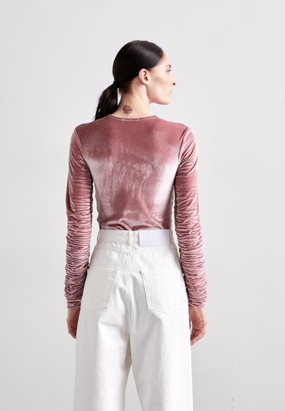 Langärmliges, pinkes Samtoberteil mit gerafften Ärmeln, kombiniert mit hoch taillierten, weißen Hosen. Die Rückansicht zeigt die glatte Textur und das taillierte Design.