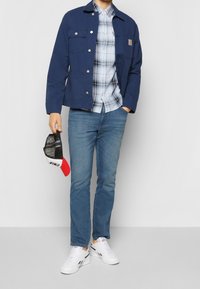 Giacca blu navy, camicia a quadri con bottoni, jeans azzurri e sneakers bianche. Berretto nero e rosso tenuto in mano. Abbigliamento casual.