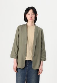 Olivengrøn, let blazer med lange ærmer, åben frontdesign og let opkrøllede manchetter, lagd over en beige top og en denimnederdel.
