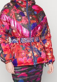 Femme portant une veste à motifs floraux colorés avec des panneaux roses brillants et une fermeture éclair bleue, associée à une jupe florale foncée.