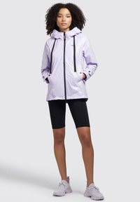 Jacket impermeabilă de culoare violet deschis cu glugă cu șnur, fermoar negru și buzunare, purtată cu șorturi negre și pantofi sport violet deschis.