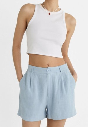 Kvinde iført hvid ærmeløs cropped top og lyseblå skræddersyede shorts med høj talje med hænderne i lommerne.