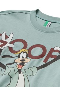 Camiseta de color verde claro con mangas cortas que presenta un gran gráfico de Goofy. Texto "GOOF" en rojo oscuro destacado en la parte delantera. Textura de algodón suave.
