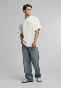 Tânăr stând cu mâinile în buzunare, purtând un tricou alb supradimensionat cu text roșu, blugi albaștri largi și adidași albi.