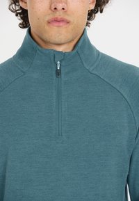 Teal fleece pullover med høj krave og halvlukning. Har en tekstureret stof og kontrastsyninger langs skuldrene.
