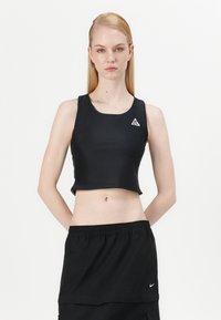 Melnas cropped stila topiņš elastīgā audumā ar apaļu kakla izgriezumu un trīsstūra logotipa plāksteri kreisajā krūtīs. Kombinēts ar melnu svārku.