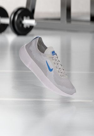 Chaussure de running Nike gris clair avec un swoosh bleu et une semelle blanche, suspendue devant du matériel de gym flou.