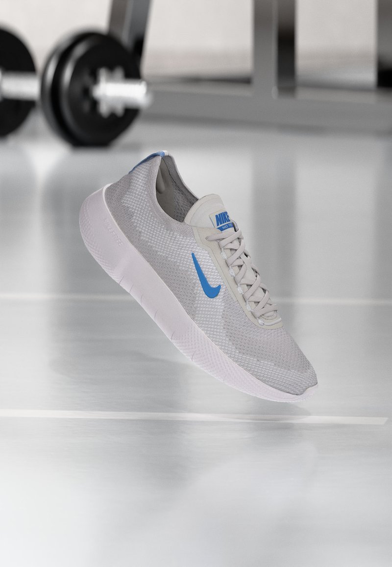 Ανοιχτό γκρι παπούτσι για τρέξιμο Nike με μπλε λογότυπο και λευκή σόλα, αιωρούμενο μπροστά από θολό εξοπλισμό γυμναστηρίου.
