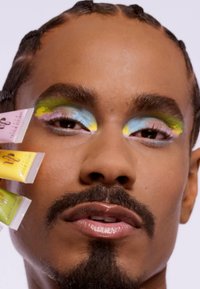 Modell med livlige pastell-sminke, med blå, rosa, gul og grønn nyanser på øyelokkene. Holder tre fargerike kosmetikk-tuber.