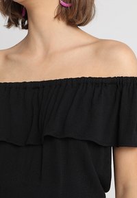 Haut noir décolleté épaules avec détail de volants froncés, tissu doux et coupe décontractée, présentant un large décolleté et une texture lisse.