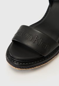 Black slide sandal made of smooth leather, featuring a wide strap embossed with "Calvin Klein" and a braided jute sole.