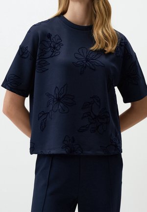 Frau trägt ein dunkelblaues Kurzarmoberteil mit bestickten Blumenmustern und passende dunkelblaue Hose.