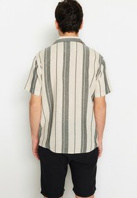 Man die met zijn rug naar de kijker staat, gekleed in een beige shirt met korte mouwen en verticale grijze strepen, en zwarte knielange shorts, tegen een witte achtergrond.