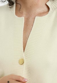 Cardigan avorio con un tessuto lavorato a maglia, scollatura a V e un singolo bottone circolare dorato sul petto. Morbidi bordi a costine sul colletto.