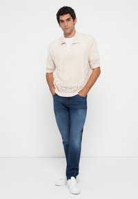 Polo shirt tricotat de un bej deschis cu guler în V, mâneci scurte, asortat cu blugi negri și adidași albi. Design și textură minimaliste.