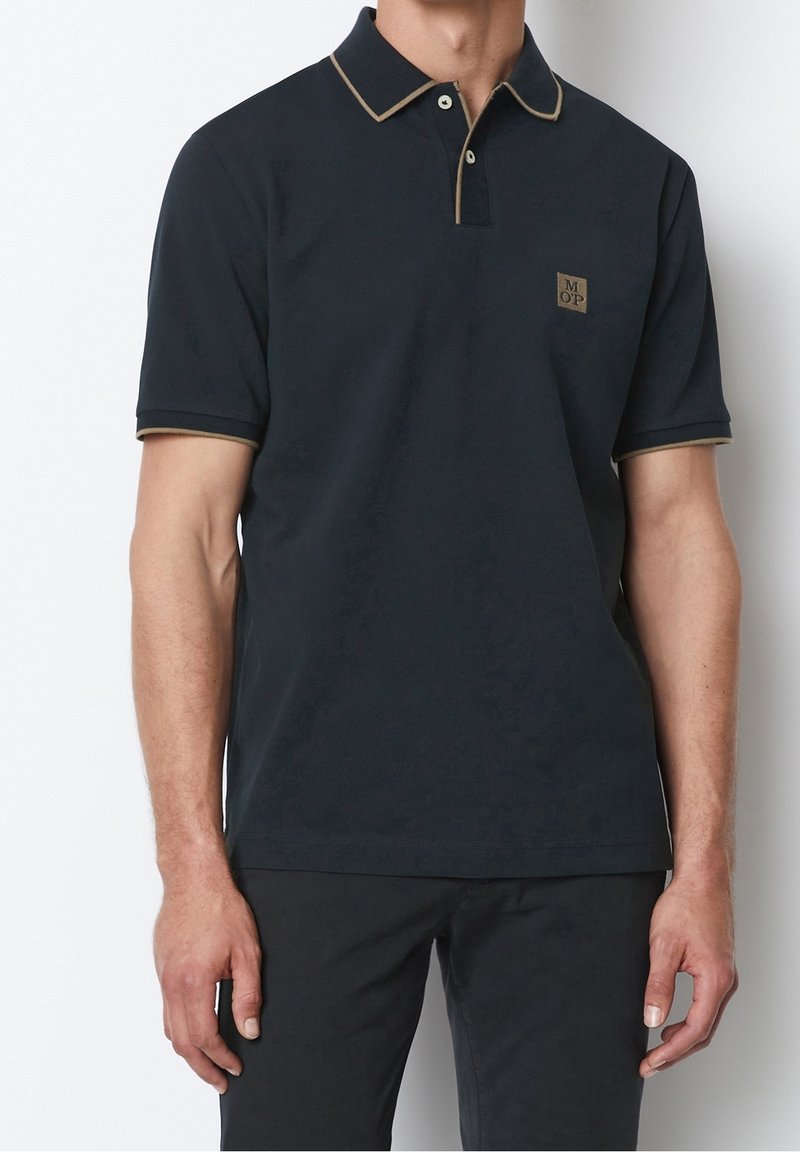 Polo-shirt bleu marine foncé à manches courtes, avec un col et des bords de manches beige en contraste, présentant un petit logo brodé sur la poitrine.