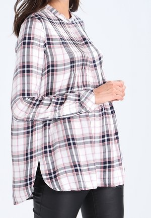 Kvinde iført en hvid ternet skjorte med lange ærmer med marineblå og røde linjer, stående med sammenknyttede hænder, kombineret med sorte bukser.