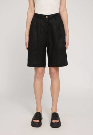 Frau trägt schwarze, knielange Shorts mit hoher Taille, Knopf und Reißverschluss, kombiniert mit schwarzen Plateausandalen zum Hineinschlüpfen und weißem ärmellosen Top.