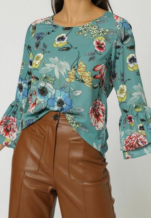 Blusa floral en azul verdoso con mangas con volantes y diversas flores en rojo, amarillo y azul. Combinada con pantalones de imitación de cuero marrón de cintura alta.