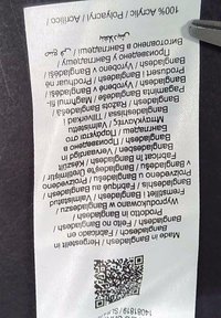 Vit tvättrådsetikett med tryckt text på flera språk, med en QR-kod, fäst på en mörk bakgrund. Texten är orienterad vertikalt.