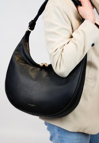 Sac à épaule en cuir noir au design courbé, matériel doré et fermeture éclair. Le sac a une texture lisse et est porté sous une veste beige.