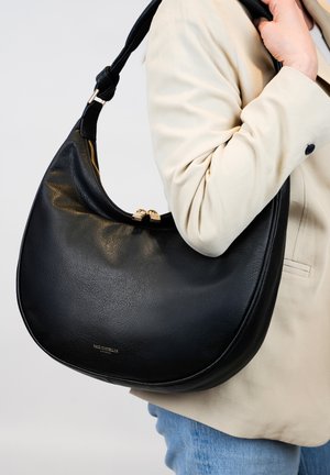 Zwarte leren schoudertas met een gebogen ontwerp, gouden hardware en een ritssluiting. De tas heeft een scherpe textuur en wordt gedragen onder een beige jas.