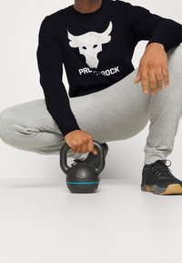 Chemise noire à manches longues avec un motif de taureau blanc, pantalon de survêtement gris, et kettlebell noir avec une accent bleu sur fond blanc.