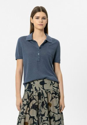 Frau trägt ein kurzärmeliges, blaues Strick-Poloshirt, das in einen hoch taillierten, gestuften Rock mit abstraktem schwarz-braunem Muster gesteckt ist.