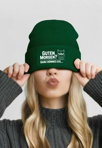 Dark green knit beanie with a flat brim, featuring white text and an owl graphic. Warm knitted texture and snug fit.