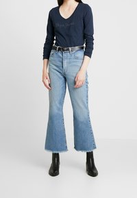 Mörkblå långärmad topp med V-ringning och broderad logotyp, parat med ljusblå utsvängda jeans med rå kant och svarta ankelboots.