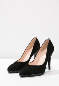 Mustat mokkasaappaat, joissa on terävä kärki ja sileä pinta. Ohut stiletto-korko on koristeltu hopeisilla niittikoristeilla pohjassa.