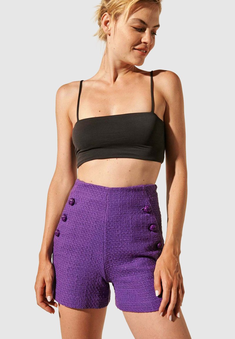 Shorts taille haute violet texturés avec de gros boutons violets sur les côtés, associés à un crop top noir. Design lisse et ajusté.
