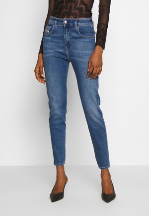 Femme portant un jean slim bleu taille moyenne, un top noir en dentelle à manches longues et des chaussures slingback noires à bout pointu, debout devant un fond uni.