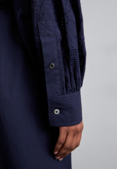 Granatowa koszula z teksturowanym haftem na rękawach, zapięciem na guziki w kontrastowym kolorze nici oraz zaprojektowanym mankietem.
