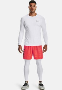 Weißes langärmliges Sportshirt, knallpinke Shorts mit schwarzem Logo, weiße Kompressionsleggings und schwarze Sportschuhe.