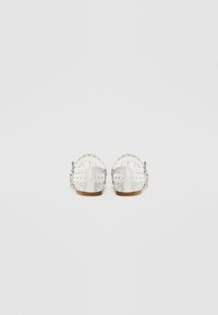 White sandals with a perforated design and metallic studs, featuring a low heel and an adjustable ankle strap. Smooth texture and rounded shape.
